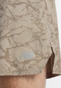 Beige athletic shorts with a speckled floral pattern, made of perforated material, featuring a reflective New Balance logo on the hem.