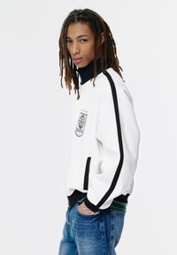 Ung person med lange dreadlocks iført en hvid jakke med sorte striber og et våbenskjold, blå jeans og et sølv kædearmbånd.