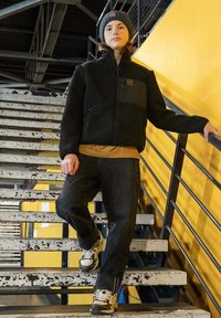 Jongere in een zwarte jas, grijze beanie en sneakers die afdaalt over versleten zwart-witte trappen met gele leuningen in een industriële omgeving.