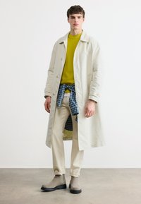 Jeune homme portant un long manteau beige, un pull jaune, un pantalon beige, des bottes grises, et une chemise à carreaux bleue nouée autour de la taille sur un fond blanc.