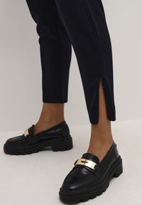 Black leather loafers with a gold metal accent, worn with navy slim-fit pants featuring a side slit at the hem.
