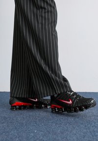 Schwarze Sportschuhe mit roten Akzenten, ausgestattet mit einem strukturierten Obermaterial, geschwungener Silhouette und einer markanten Sohle mit luftgepolsterten Blasen.