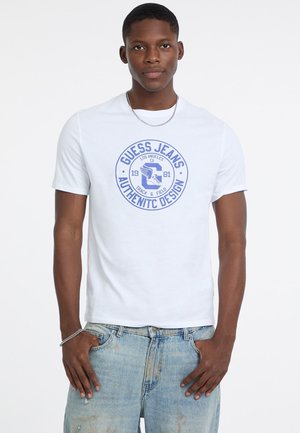 Młody mężczyzna w białej koszulce Guess Jeans z niebieskim logo i jasnoniebieskich zniszczonych dżinsach, stojący z rękami w kieszeniach.