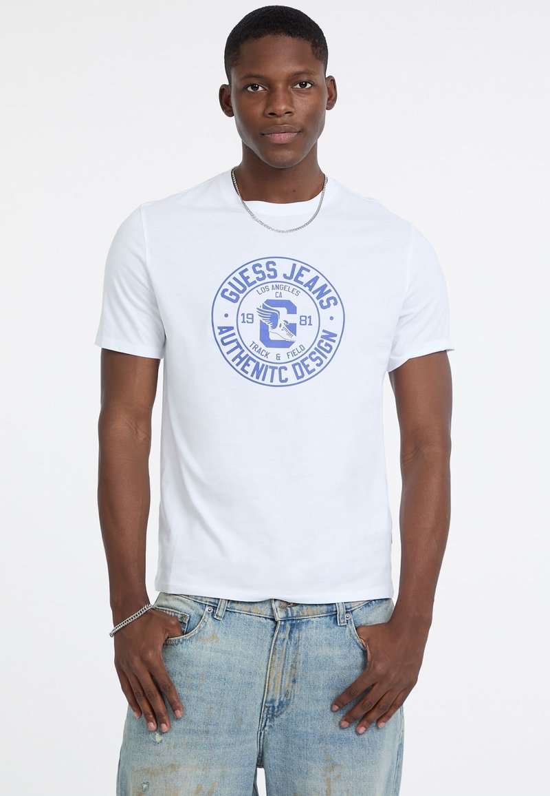 Młody mężczyzna w białej koszulce Guess Jeans z niebieskim logo i jasnoniebieskich zniszczonych dżinsach, stojący z rękami w kieszeniach.