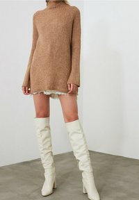 Donna che indossa un maglione a collo alto rigato color cammello, una mini gonna bianca sfilacciata e stivali bianchi in pelle con tacco alti fino al ginocchio, in piedi all'interno.