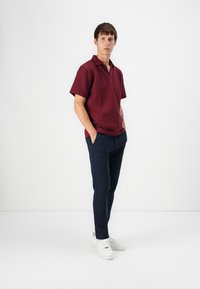 Short-sleeve polo shirt in burgundy with textured knit pattern, paired with navy slim-fit trousers and white sneakers. Model poses with hands in pockets.