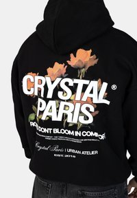 Černá mikina s kapucí, která na sobě má květinovou grafiku a bílé nápisy "CRYSTAL PARIS" a "RŮŽE NEKVETOU V POHODLÍ." Vyrobena z měkké bavlněné směsi.