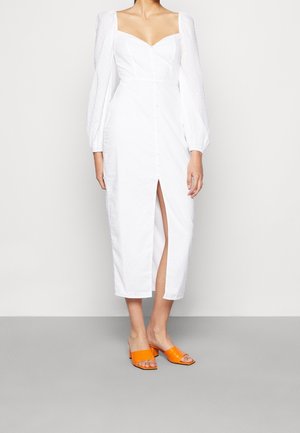 Robe en lin blanc avec manches bouffantes, décolleté en cœur et boutonnage devant. Comprend une fente haute sur la cuisse et est associée à des sandales à talons orange.