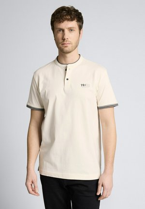 Mann mit kurzem lockigem Haar, der ein beigefarbenes Henley-Shirt mit kurzen Ärmeln, schwarzem Rand und dem Text "19/62" auf der Brust trägt, steht vor einem einfarbigen Hintergrund.