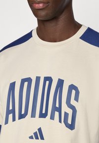 Bež trenirka z mornarsko modrimi raglan rokavi, na sprednji strani velika modra napis "ADIDAS" in logotip, izdelana iz mehke bombažne tkanine.