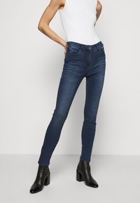 Jeans ajustados de color azul oscuro hechos de denim, con un diseño de bolsillo frontal y una bragueta con cremallera. Combinados con botas de tobillo negras y una camiseta sin mangas blanca.