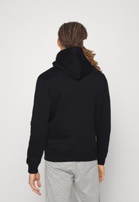 Felpa nera con cappuccio e cordino, caratterizzata da un tessuto testurizzato e polsini a coste. Indossata con pantaloni della tuta grigi, vista da dietro.