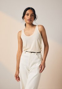 Débardeur beige clair côtelé avec larges emmanchures et encolure dégagée, associé à un pantalon taille haute crème et une ceinture noire.