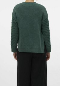 Zielony sweter z dzianiny z żebrowanym kołnierzem i teksturowanymi rękawami w diamentowy wzór. Szeroki krój z bocznymi rozcięciami i gładkim wykończeniem. Czarne spodnie.