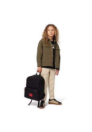 Sac à dos noir à motifs avec détail de logo rouge, porté par une personne en veste olive, t-shirt noir, pantalon cargo beige et baskets noires.