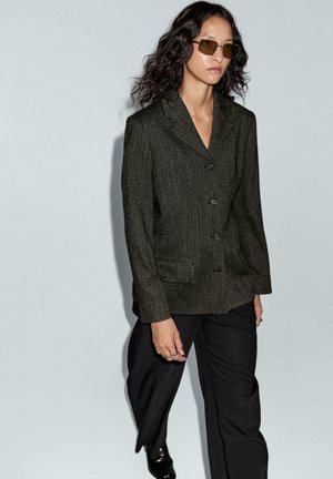 Blazer en tissu texturé vert foncé avec un col cranté, fermeture à deux boutons et poches avant, porté avec un pantalon noir ample.