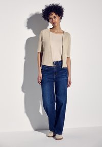 Beige gestrickte, cropped Strickjacke über einem hellen Oberteil, kombiniert mit hochtaillierten blauen Jeans und beigen Schuhen. Glatte Textur und lockere Passform.