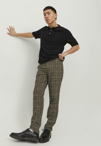 Sort polo shirt, brune ternede bukser med blå og gule accenter og sorte snørestøvler. Stående pose foran en ensfarvet baggrund.