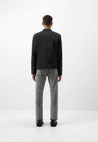 Zwarte jas met een minimalistisch ontwerp, gecombineerd met grijze straight-leg broek en zwarte schoenen, van achteren gezien op een witte achtergrond.