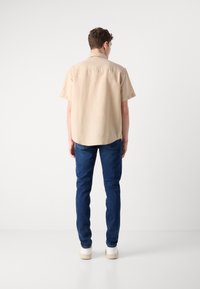 Camicia beige a maniche corte con orlo curvo, abbinata a jeans blu scuro e sneakers beige, vista da dietro.