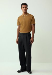 Camisa polo marrón con cuello blanco a rayas, de manga corta, combinada con pantalones negros con pliegues y mocasines negros. Diseño y ajuste simples.
