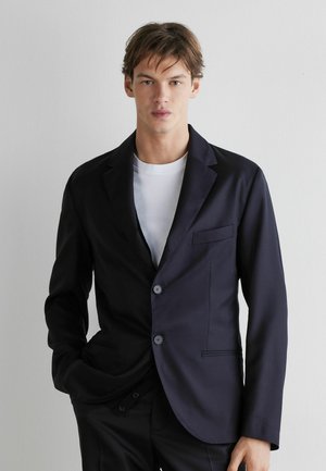 Junger Mann trägt einen dunkelblauen Blazer über einem weißen T-Shirt, steht vor einer schlichten hellgrauen Wand mit einer Hand in der Tasche.