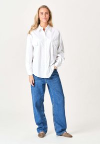 Ung kvinde med langt blondt hår iført en løs hvid skjorte med knapper, blå vide jeans og taupe flade sko, stående med den ene hånd i lommen.