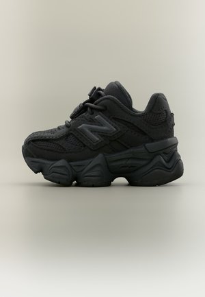 Baskets épaisses noires New Balance avec semelle épaisse, panneaux en mesh et daim, et grand logo "N" sur le côté, montrées de profil sur fond beige.