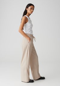 Pantalons beige à jambes larges avec une taille à cordon, dotés de poches latérales et d'une texture lisse. Associés à un débardeur blanc ajusté.