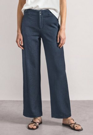 Femme portant un pantalon droit bleu foncé, un haut blanc et des sandales noires avec des brides dorées, debout sur un sol gris.