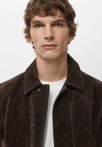 Smeđa jakna od brušene kože s patentnim zatvaračem sprijeda i kopčama na pritisak, s ovratnikom od korduroy tkanine i glatkom teksturom. Bijela majica ispod.