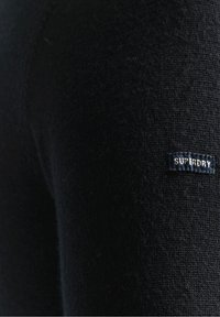 Schwarzer Stoff mit einer strukturierten Oberfläche. Verfügt über ein kleines blaues Etikett mit weißem Text, der "SUPERDRY" liest. Nahaufnahme des Materials.