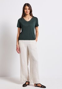 Camiseta de cuello en V de manga corta en verde oscuro con acentos dorados, combinada con pantalones de lino de pierna ancha en color blanco roto y sandalias tejidas negras.