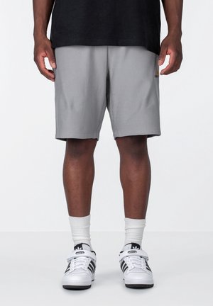 Personne portant un short gris, des chaussettes blanches courtes et des baskets Adidas blanches avec des rayures noires sur un fond uni.