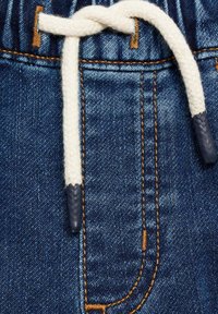 Uvećani prikaz denimske tkanine u plavoj nijansi, zlatnim šavovima i bijelom vezicom vezanom u čvor s tamnim vrhovima.