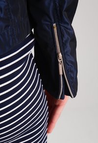 Navy nylon jacket with a zippered cuff, featuring a sleek design and smooth texture. Paired with a navy-and-white striped dress.