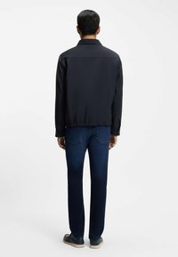 Man standing facing away, wearing a black jacket, dark blue jeans, and black shoes with white soles against a plain white background.