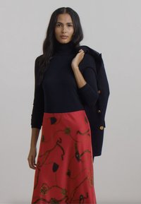 Svart turtleneck genser kombinert med et rødt skjørt med gulldetaljer og en svart blazer drapert over skulderen. Glatte stoffstrukturer.