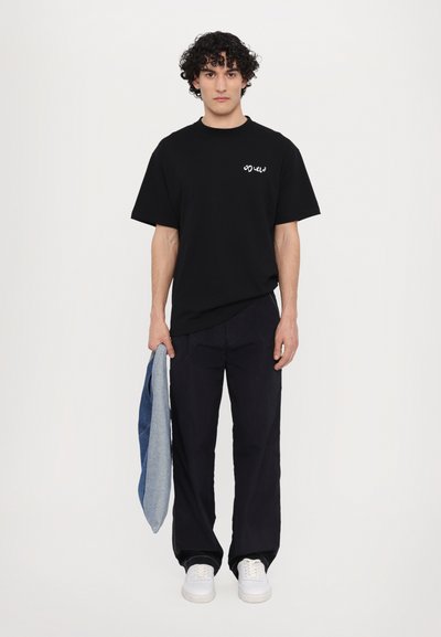 T-shirt nera con un piccolo logo bianco, abbinata a pantaloni blu navy e sneakers bianche. Tiene in mano una giacca di jeans. Design semplice, vestibilità rilassata.