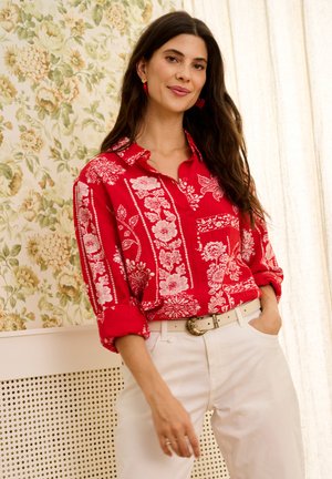 Žena s dlhými tmavými vlasmi, oblečená v červenej kvetinovej košeli na gombíky a bielych nohaviciach, stojaca s jednou rukou v vrecku pred kvetinovou tapetou.