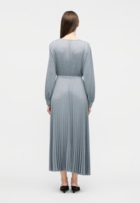 Γυναίκα με μακριά καστανά μαλλιά ντυμένη με ασημί-γκρι μίντι πλισέ φόρεμα με μακριά μανίκια και μαύρα mules με τακούνι, που στέκεται γυρισμένη προς τα πίσω.