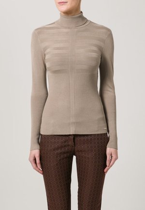 Femme portant un pull à col roulé côtelé beige et un pantalon marron à motifs, debout devant un fond clair uni.