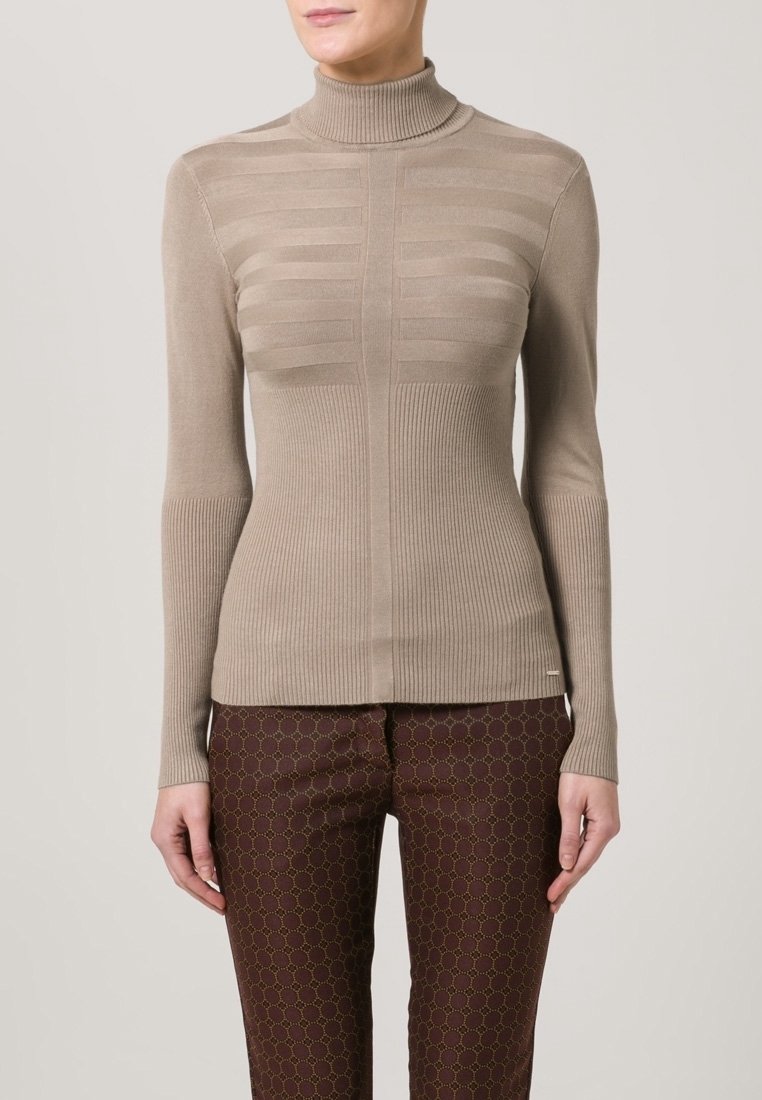 Femme portant un pull à col roulé côtelé beige et un pantalon marron à motifs, debout devant un fond clair uni.