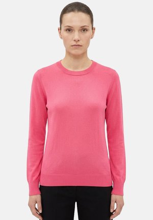 Donna con i capelli raccolti che indossa un maglione a maniche lunghe di colore rosa acceso e pantaloni neri, in piedi davanti a uno sfondo chiaro e uniforme.