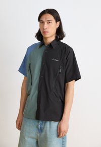 Short-sleeve shirt in black and teal with a partial zip, featuring contrasting fabric sections and a logo. Lightweight, smooth texture.
