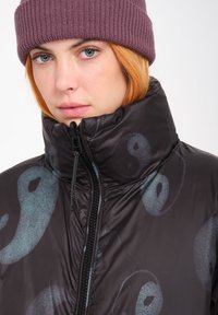 Czarna kurtka puchowa z wysokim kołnierzem, z subtelnym wzorem yin-yang w odcieniu turkusowym. Zamek błyskawiczny oraz teksturowana czapka beanie.