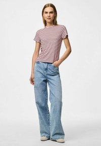 Rozā un baltā svītrainā t-kreklā ar īsām piedurknēm, apvienots ar gaiši ziliem, brīvi pieguļošiem džinsiem. Modelis stāv ar vienu roku kabatā.