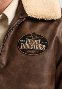 Chaqueta de piel marrón con un cuello de borreguillo crema. Presenta un parche circular con la inscripción "Petrol Industries" en bordado negro y dorado.