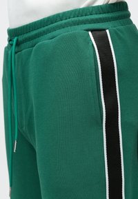 Pantaloni de trening verzi cu talie elastică, șnur verde, buzunar lateral și detalii cu dungi verticale alb-negru.