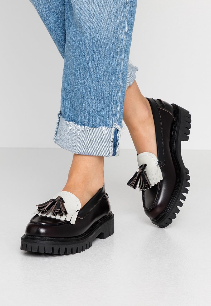 Mocassins en cuir noir avec un accent en cuir blanc et des pampilles, dotés d'une semelle épaisse et robuste noire et d'une texture lisse et brillante.